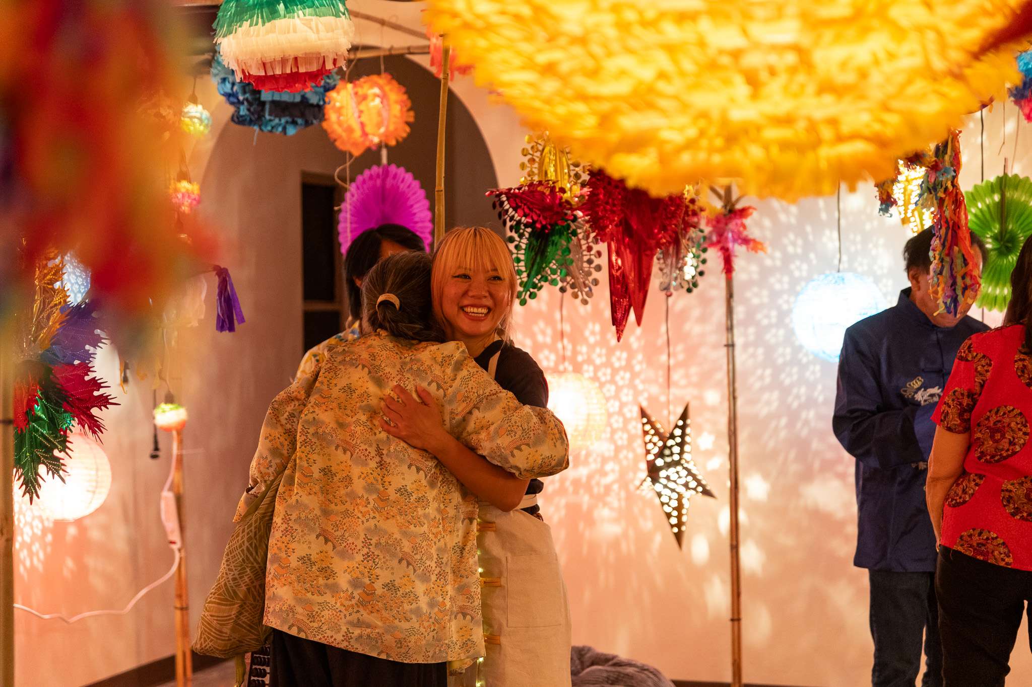 A Chinese American chef hugs an elder under illuminated pinatas.