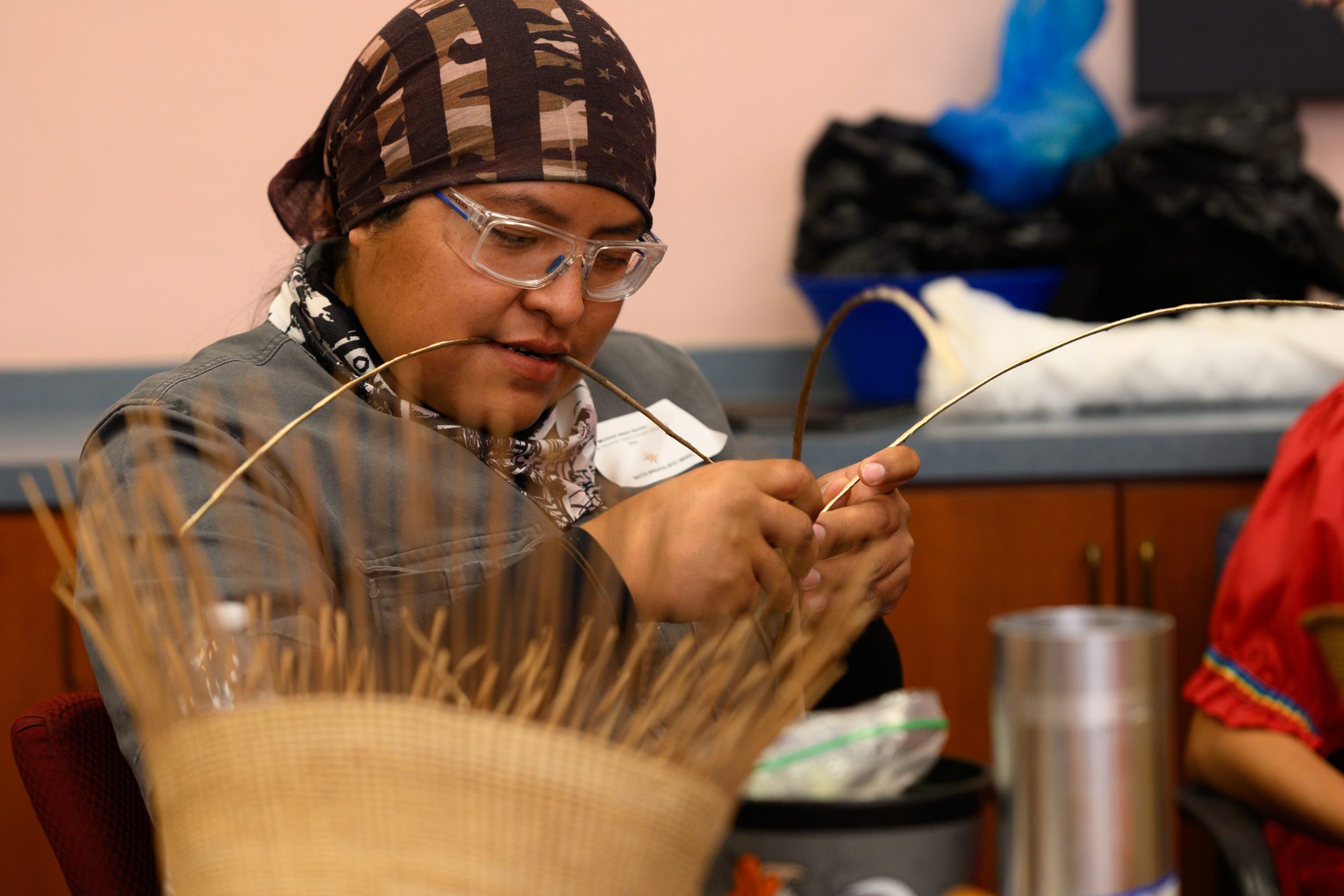 A young Native person in a headband is weaving together natural fibers with a piece in their mouth.