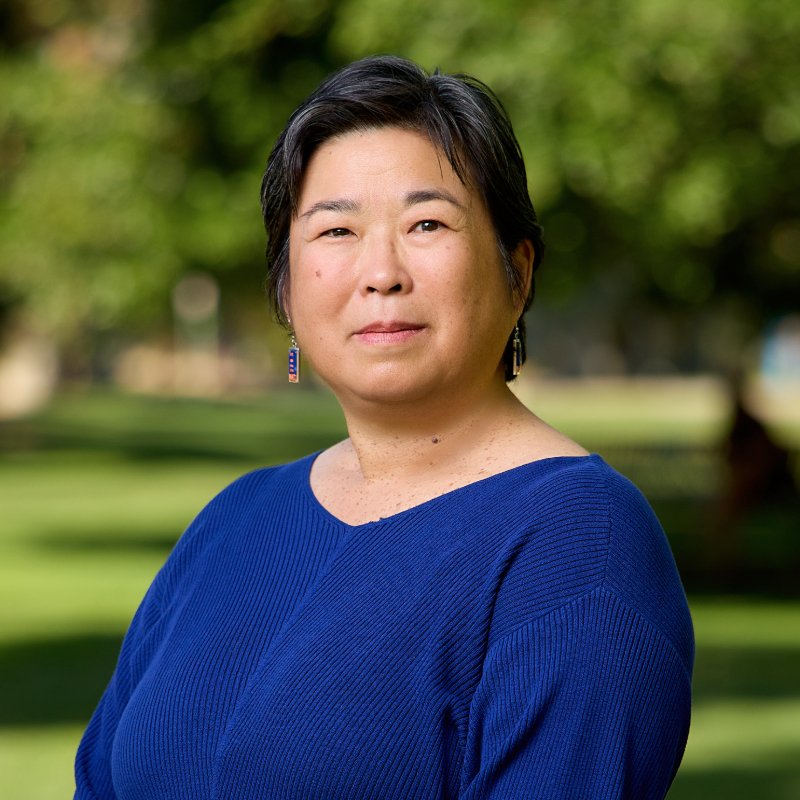 A smiling Japanese American woman of Okinawan descent with dark hair and light brown skin wearing a blue blouse.