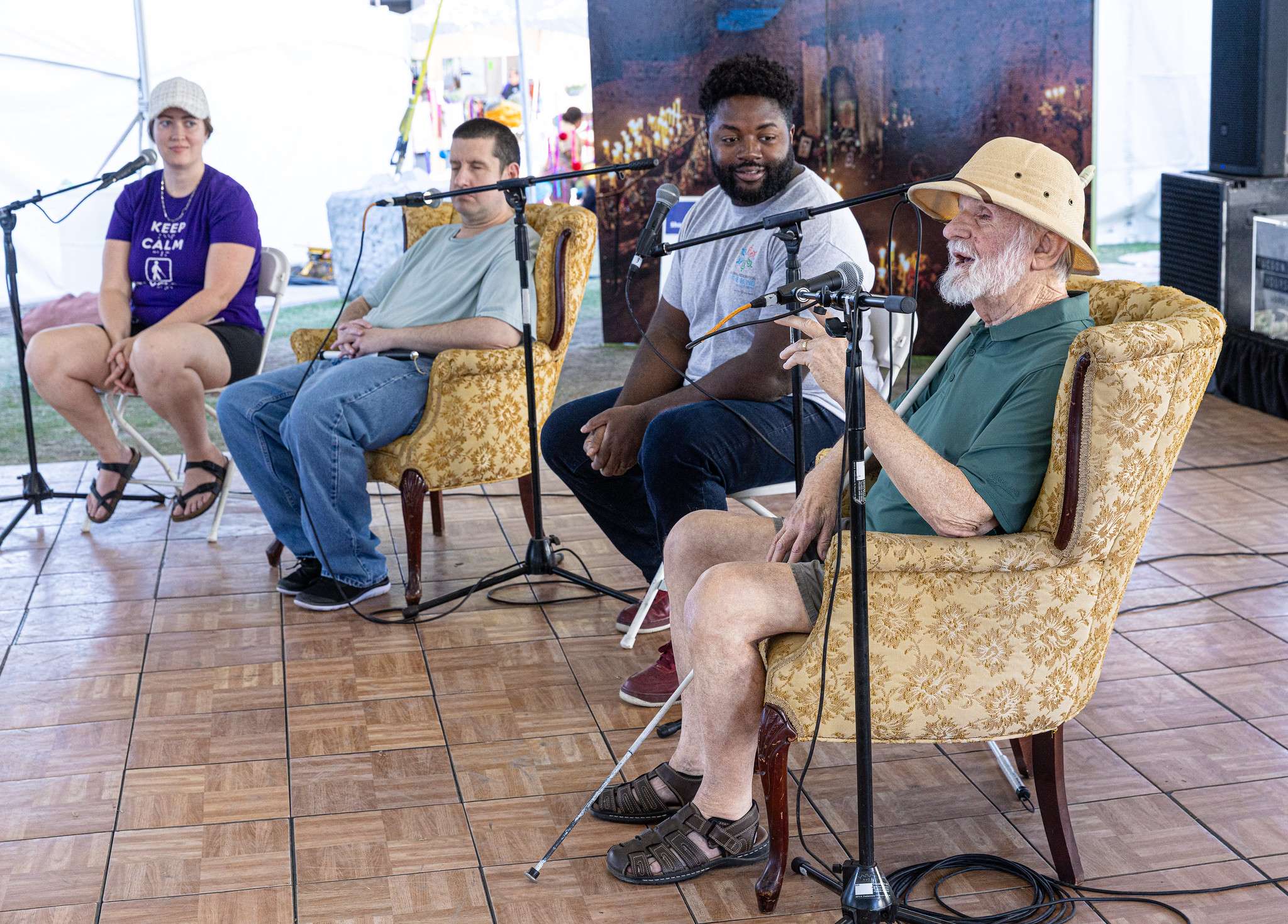 Four people sitting in chairs with mic stands in front of them. An older man with a whit beard, explorer hat, and white cane is speaking into a mic.