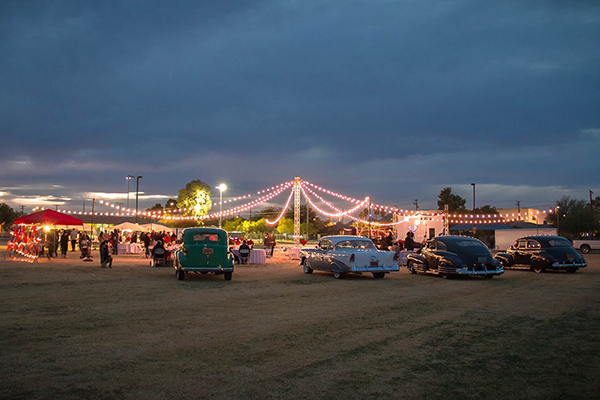 Old school cars parked in front a a circle of lights. People are walking under the lights.
