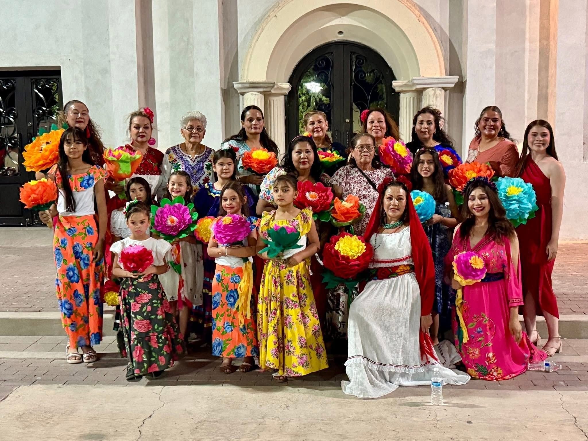 An intergenerational group of about 25 women and girls in brightly colored dresses and skirts with flowers pose in three rows. Many are holding large, brightly colored paper flowers.