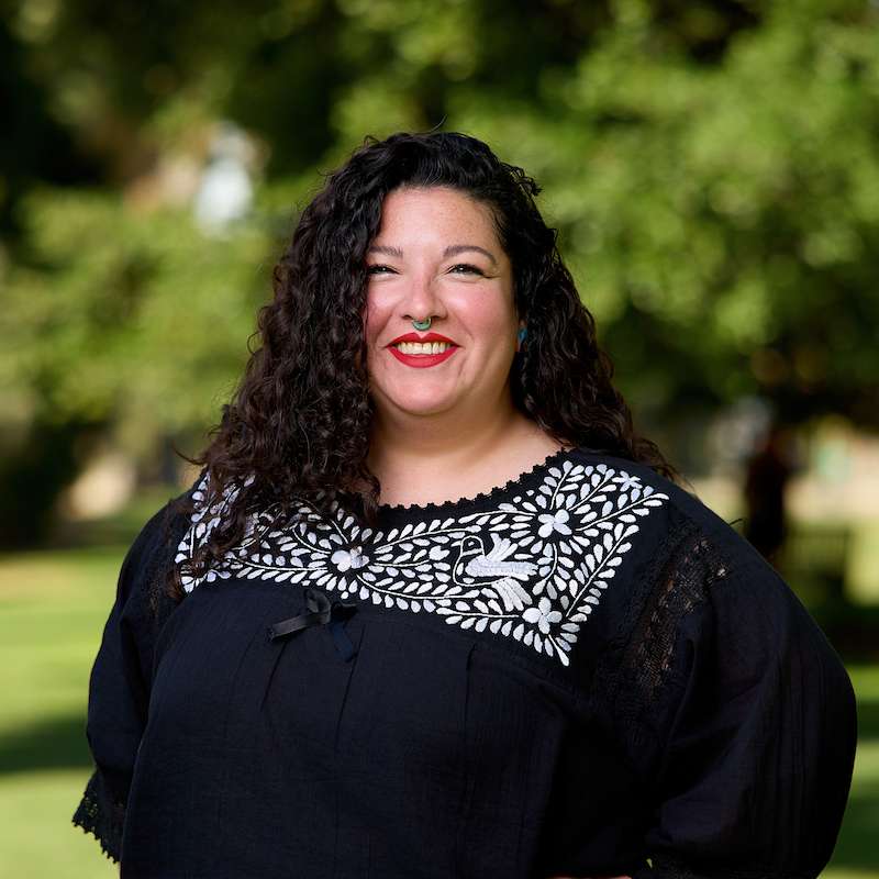 A head shot of a smiling woman with long curly black hair, red lipstick, and a black top with white flowers.
