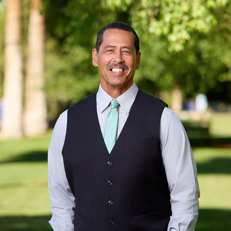 A head shot of a man with medium brown skin, a mustache and dark hair wearing a dark vest with a light blue shirt and tie underneath.