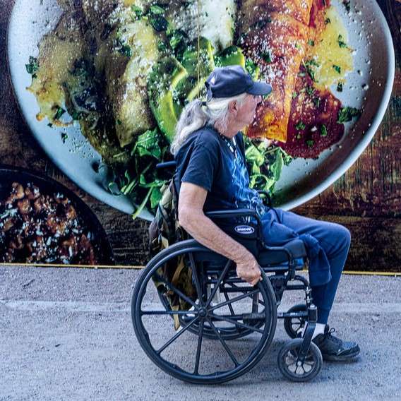 An older person with light skin and gray hair in a ponytail is in a wheelchair in front of a plate of green and red enchiladas.