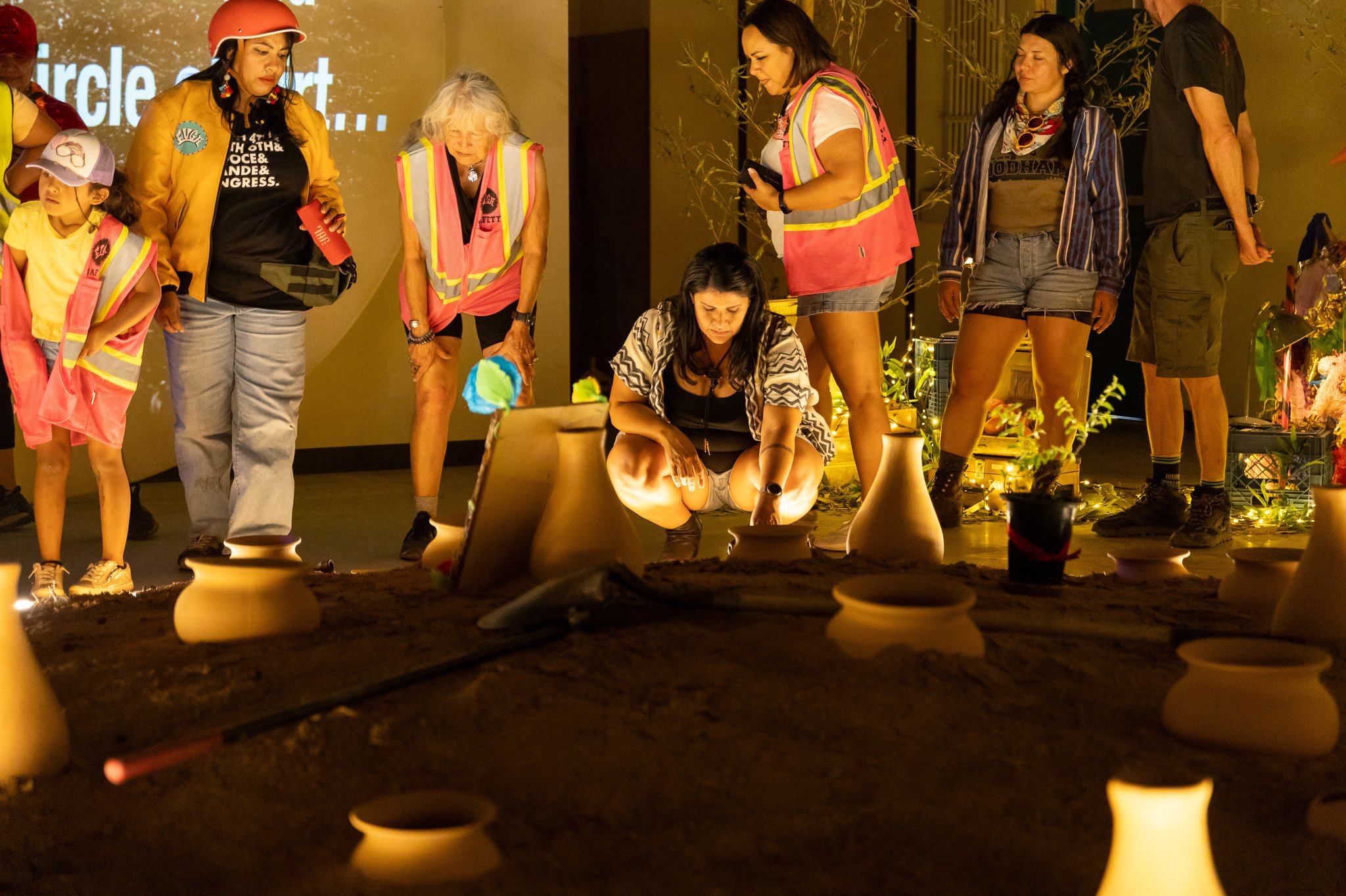 A group of people in bicycle gear and reflective vests gather around part of a circular altar. One person is crouching down with their hand in the dirt pile with clay pots.