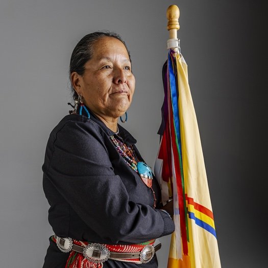 A Diné woman holding a Navajo flag is wearing a black shirt with jewelry and a belt.