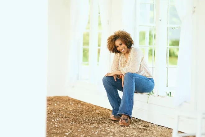 Woman wearing blue jeans and a white blouse smiles from sitting at a window.