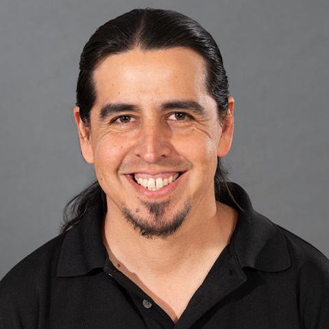 A smiling Chicano man with long dark black hair pulled back. He is wearing a black collared shirt.